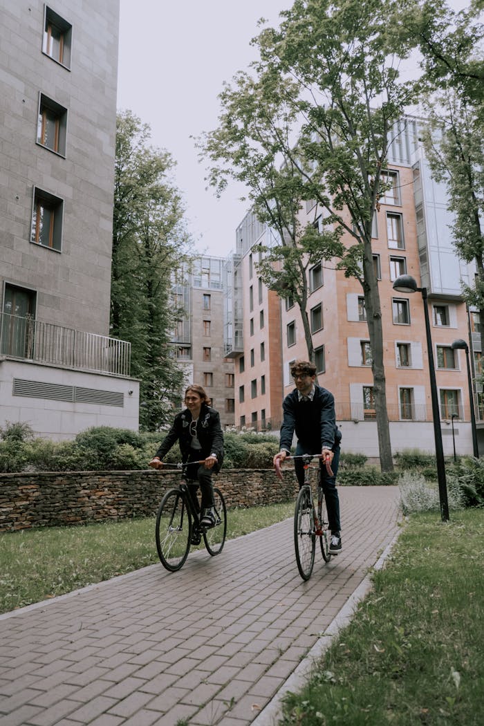 Two friends cycling in an urban setting with modern apartments around, enjoying recreation outdoors.